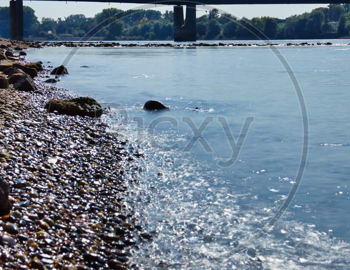 Blue Water In The Rhein River Hitting The Rocks On Shore In Germany On A Summer Day.