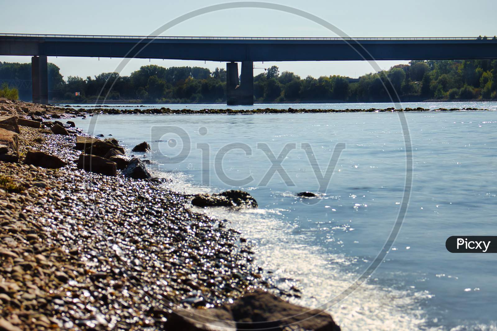 Water Hitting The Rocky Shore With A Bridge In The Background On A Warm Summer Day In Germany.