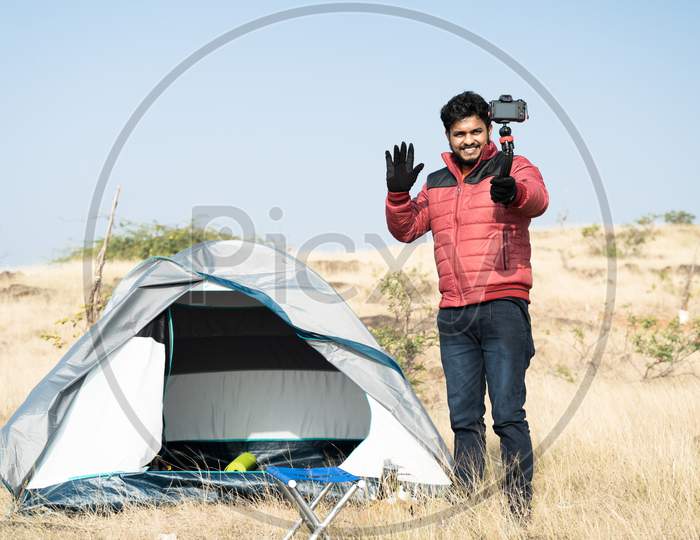 Wide Angle Shot Of Young Traveller Busy Talking With Camera On Top Of Mountain In Front Of Camping Tent - Concept Of Travel Vlogger, Blogger Or Influencer Recording Video During Hiking.