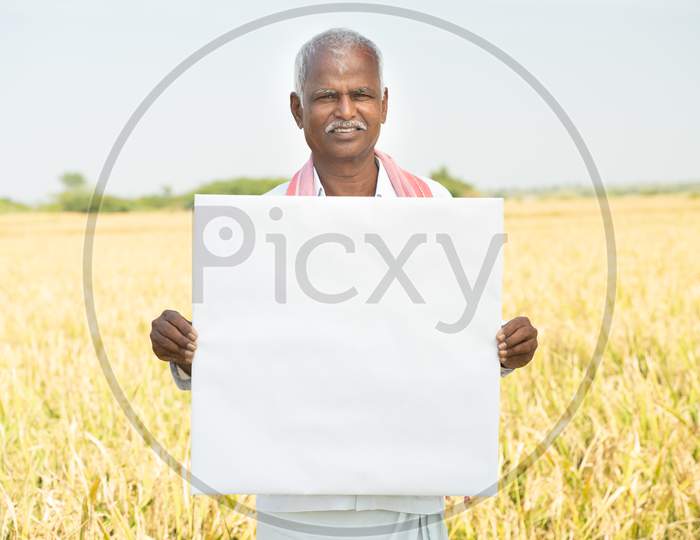Smiling Indian Farmer Holding White Card Board Or Empty Banner Infornt Of The Agriculture Farmland.