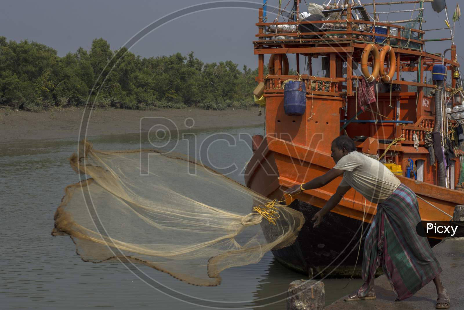 A Fisherman Throwing His Fishing Net At Water For Catching Fish, His Boat Is In Background.