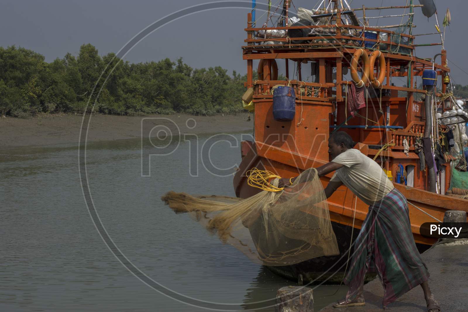 Image of A Fisherman Throwing His Fishing Net At Water For Catching Fish,  His Boat Is In Background.-SF973302-Picxy