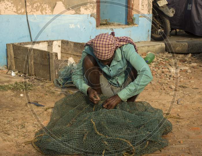 A Fisherman Stitching His Fishing Net Sitting In Front Of His House.