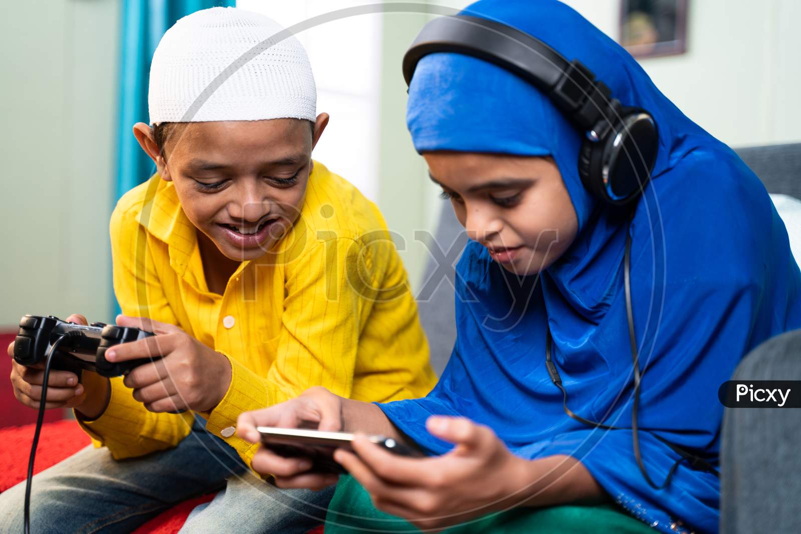 Concept Of Kids Busy With Modern Day Technology Lifestyle And Distraction - Two Muslim Siblings, Brother Playing Game Using Joystick And Sister Playing Games On Mobile Phone With Headphones At Home.