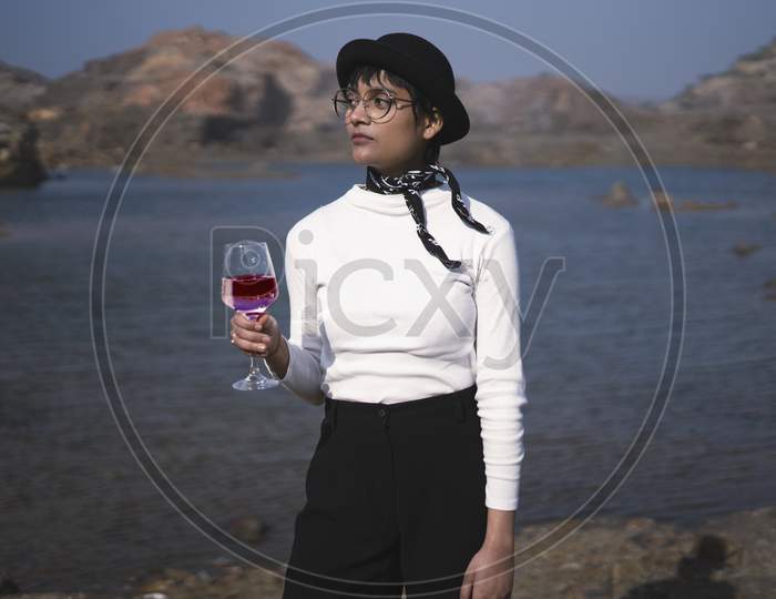 Young Beautiful Girl Standing Near A Lake With A Beautiful Scenery In The Background, Holding A Wine Glass In Her Hands And Wearing A Black Hat. Life Style Concept.