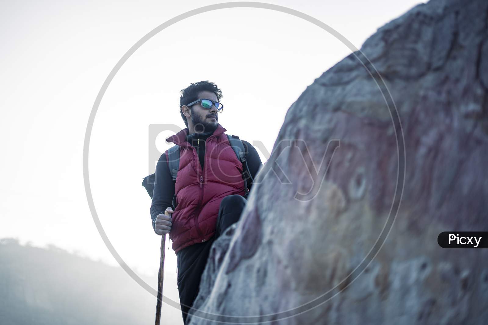 Wide Angled Shot Of A Indian Traveler Hiking Up The Mountain, Shot At A Shallow Depth Of Field From A Rock. Trekking With A Backpack And With Red Snow Jacket.