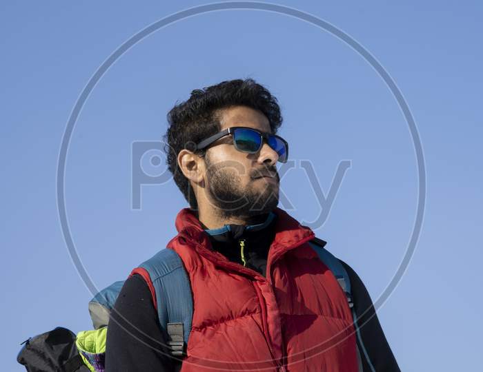 Portrait Of An Young Indian Traveler Ready For Hiking Wearing Red Snow Jacket And A Backpack. Freedom And Activeness Concept.