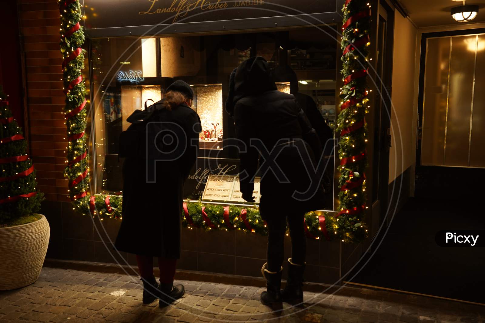 Two Female Pedestrians Standing In Front Of A Store Window In Dark Evening. Focus On Window.