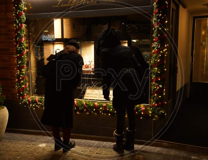 Two Female Pedestrians Standing In Front Of A Store Window In Dark Evening. Focus On Window.