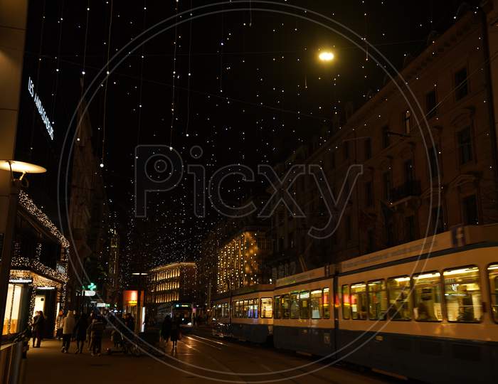 Pedestrian And Tram In The Evening, Under Hanging Lamps Called Lucy, At Bahnhofstrasse At Christmastime In Time Of Corona Virus.