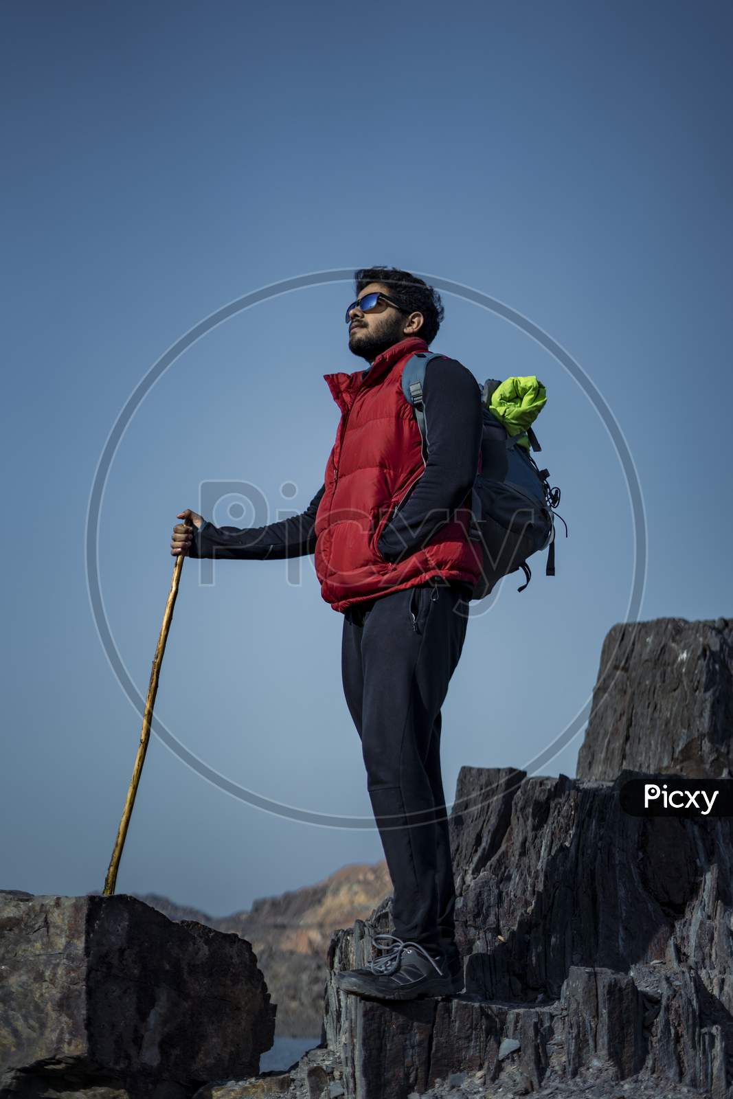 Young Indian Traveler Hiking Up The Mountain With A Backpack And A Stick, With A Beautiful Lake View In The Background. Sports And Freedom Concept.