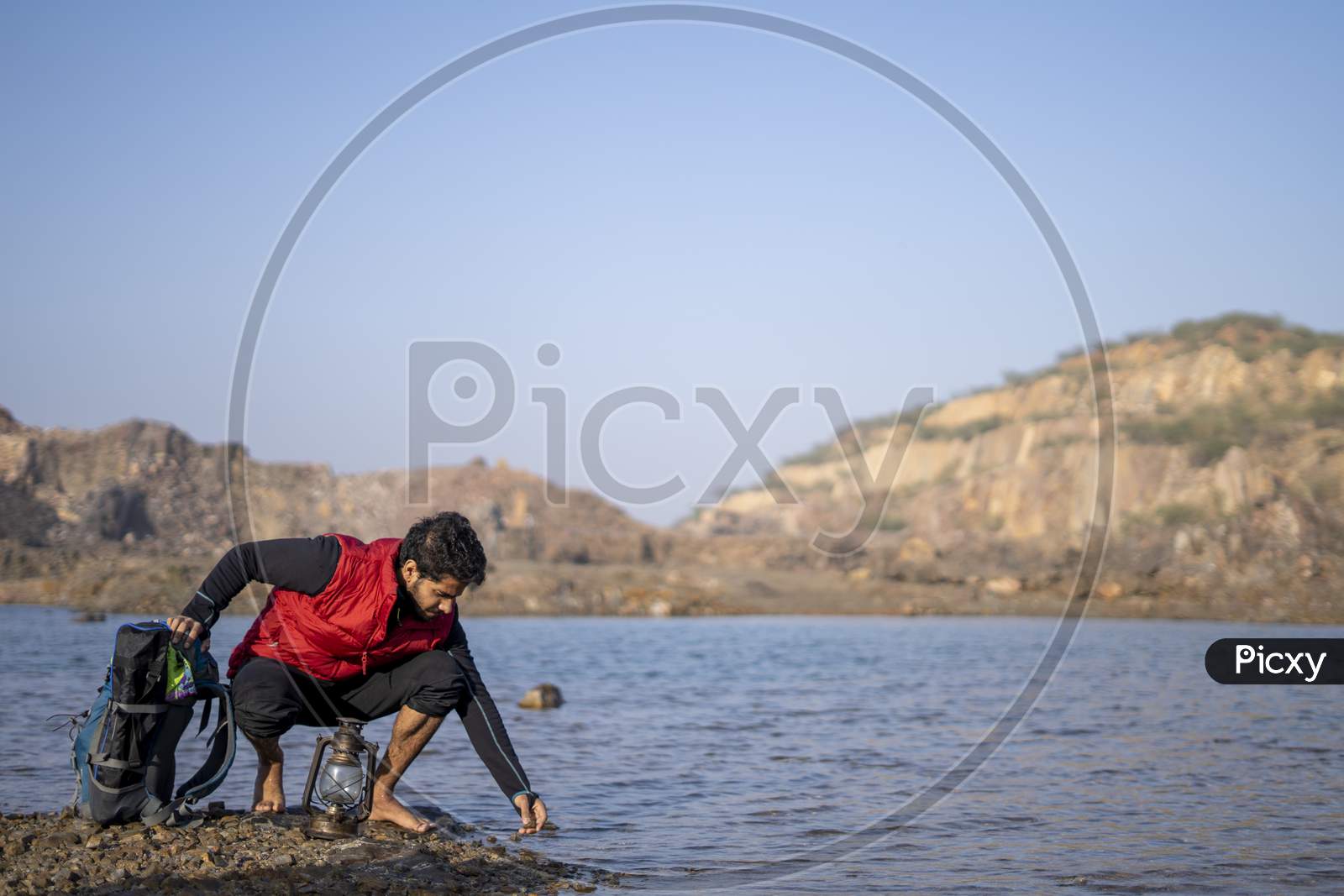 Young Indian Traveler Getting Ready For Camping In The Mountains, Sitting Near A Lake With His Backpack. Getting Water For Camping. Freedom And Adventure Concept.