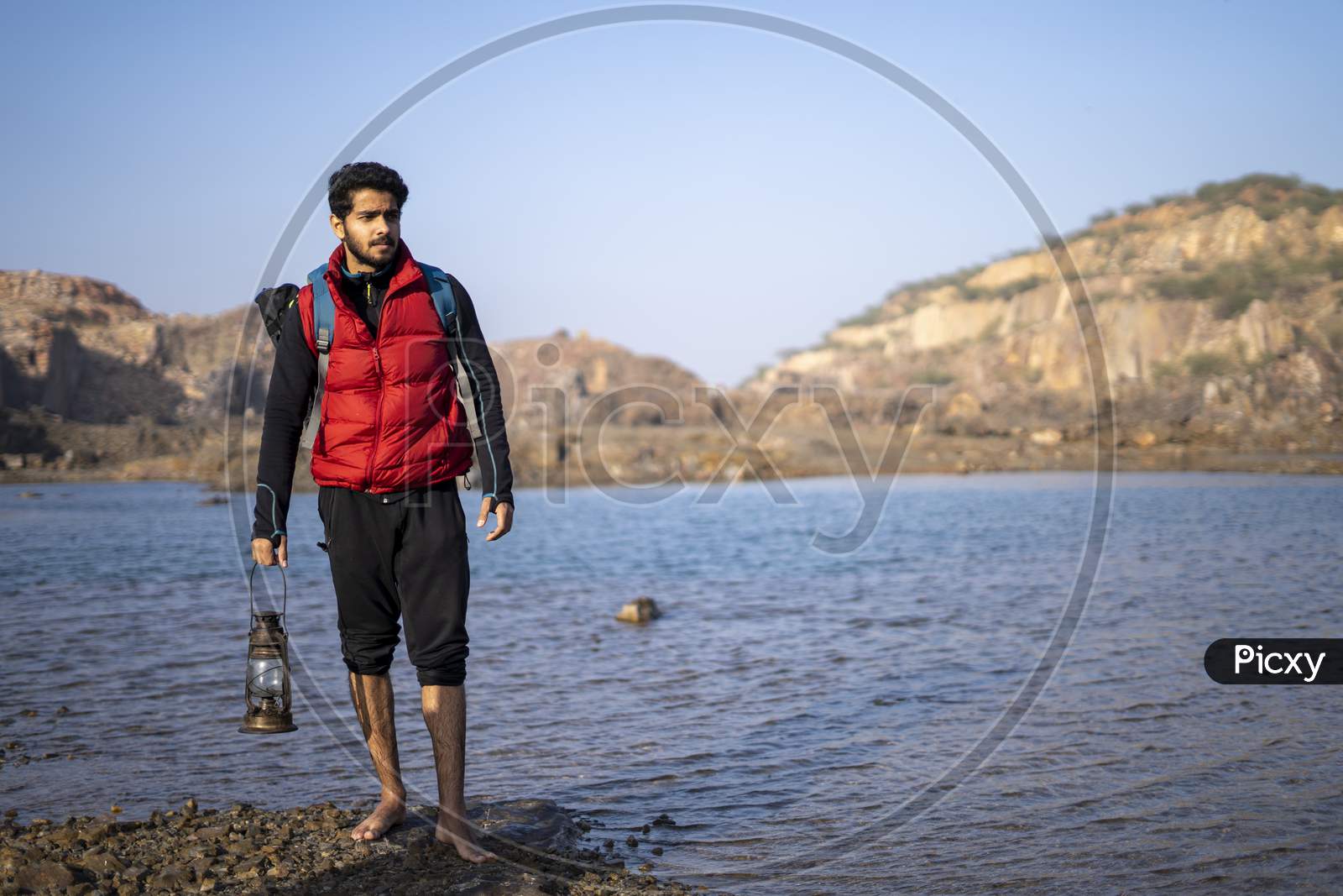 Young Indian Traveler Standing Near A Lake In The Mountains With A Lantern, Getting Ready For Camping In The Night.