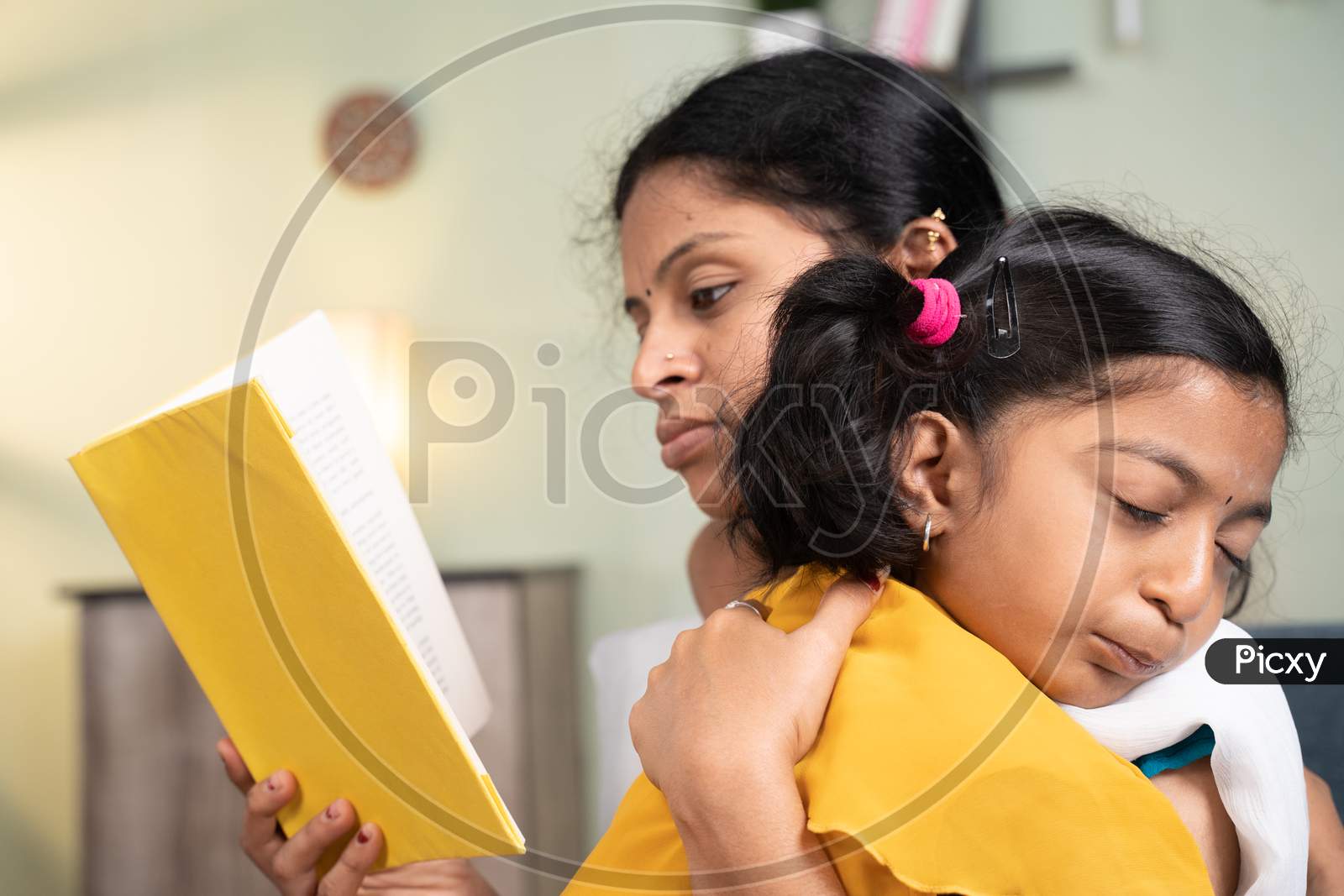 Girl Kid Sleeping On Mother'S Shoulder While Mother Busy Reading Book And Making Her Kid Sleep At Home.