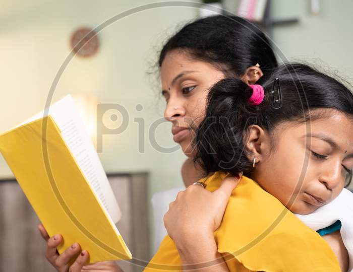 Girl Kid Sleeping On Mother'S Shoulder While Mother Busy Reading Book And Making Her Kid Sleep At Home.