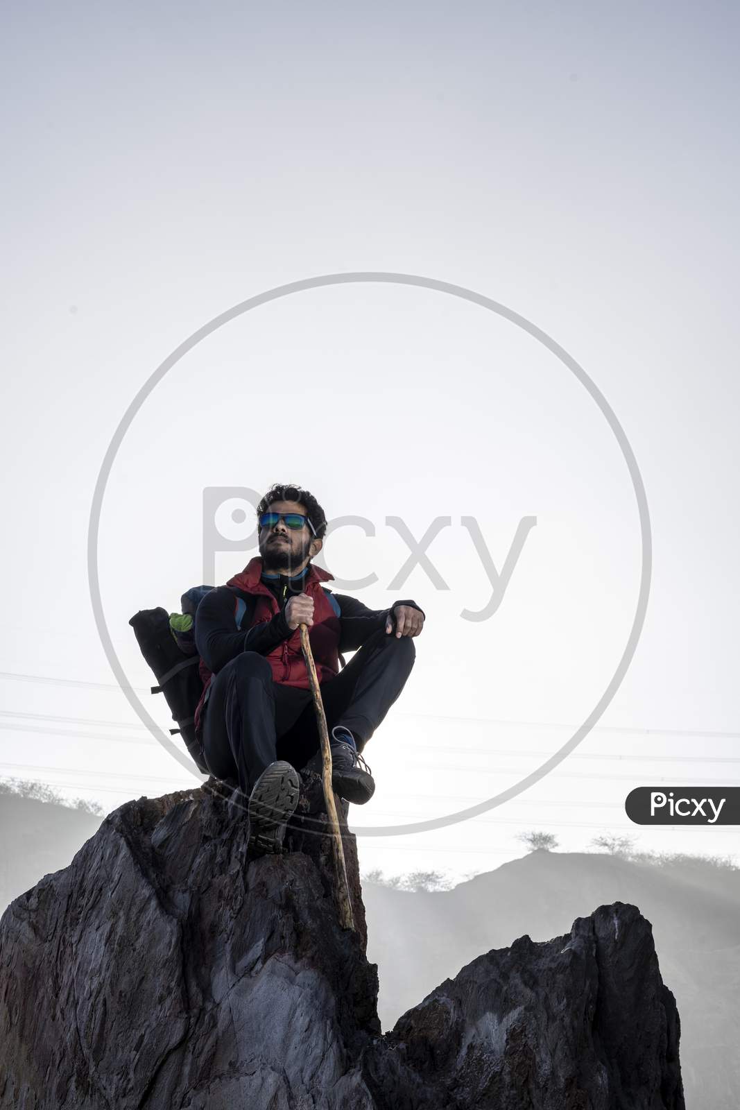 Young Indian Mountaineer Sitting On The Top Of The Mountain On A Cliff With A Backpack And A Stick, Enjoying The View From The Top Of The Mountain, Captured During Sunset.