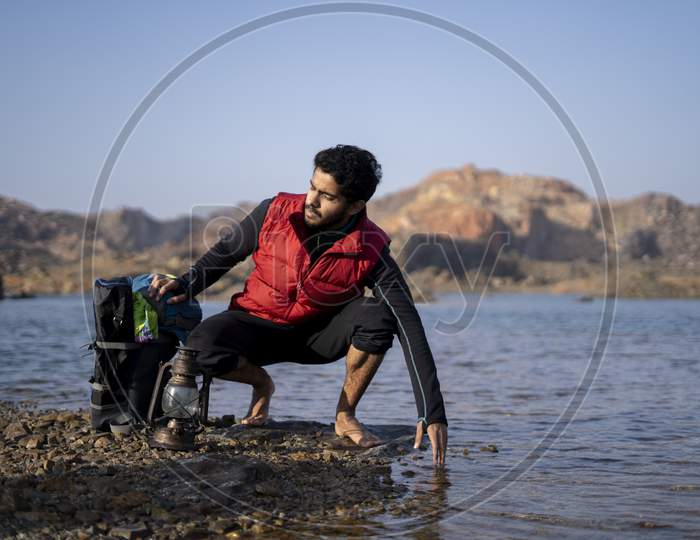 Young Indian Traveler Getting Ready For Camping In The Mountains, Sitting Near A Lake With His Backpack. Getting Water For Camping. Freedom And Adventure Concept.