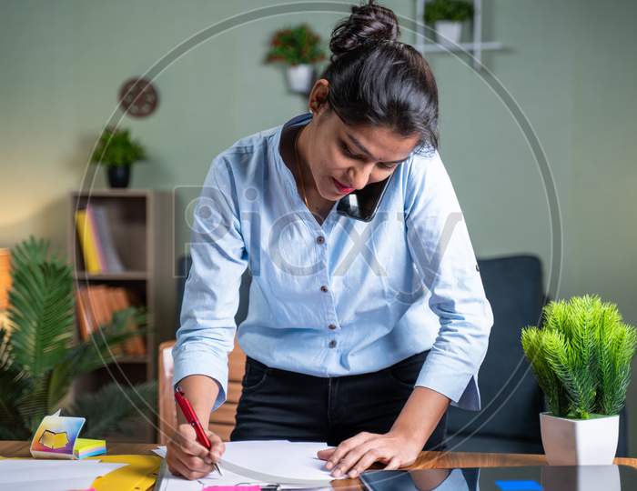 Standing Young Indian Business Woman Busy Making Notes While Talking On Mobile Phone In Front Of Working Desk - Concept Of Business Call, Talking With Clients Noting Down Tasks.