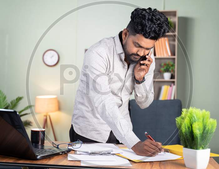 Standing Young Man Busy Making Notes While Talking On Mobile Phone In Front Of Working Desk - Concept Of Business Call, Talking With Clients Noting Down Tasks.