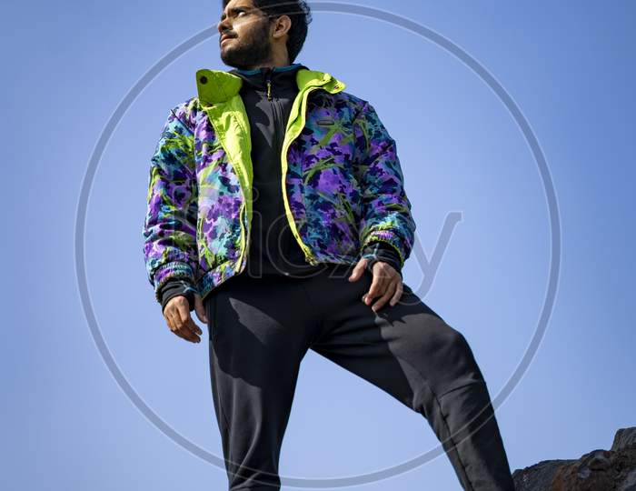 Young Indian Boy Standing On The Top Of Cliff Wearing A Snow Jacket And Track Pants. Enjoying And Freedom Concept.