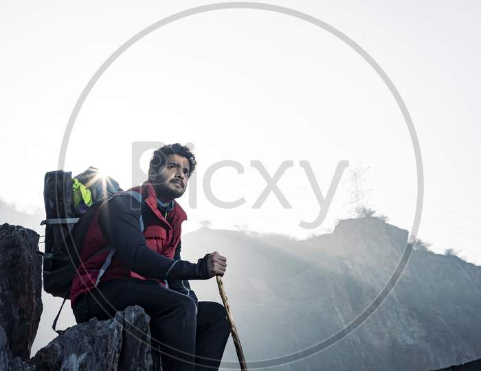 Young Indian Mountaineer Sitting On The Top Of The Mountain On A Cliff With A Backpack And A Stick, Enjoying The View From The Top Of The Mountain, Captured During Sunset.