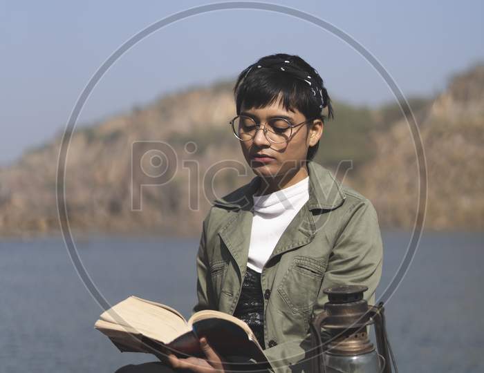 Young Beautiful Studious Girl Reading Books Near A Lake In The Mountains While Sitting On A Rock, With A Head Gear On Her Head. Travel And Education Concept.