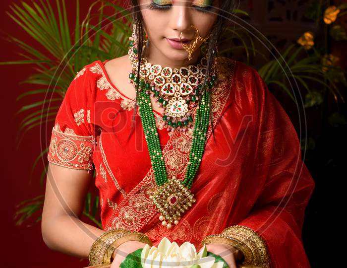 Portrait Of Very Beautiful Young Indian Bride In Luxurious Bridal Costume With Makeup And Heavy Jewellery Holding A Lotus In Studio Lighting Indoor. Lifestyle Wedding Fashion.