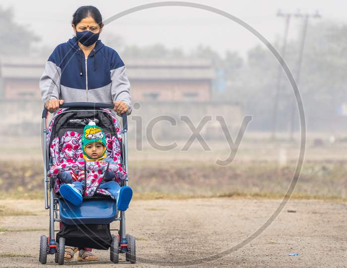 An Elderly Indian Lady Wearing Black Face Mask Pushing An Indian Baby In Winter Wear In A Stroller Perambulator On A Road With Copy Space And Background Blur
