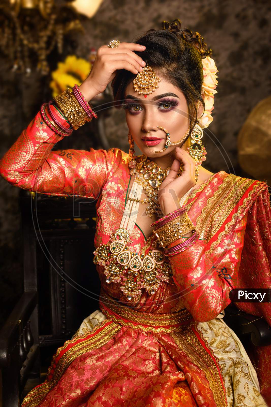 Magnificent Young Indian Bride In Luxurious Bridal Costume With Makeup And Heavy Jewellery Is Sitting In A Chair In With Classic Vintage Interior In Studio Lighting. Wedding Fashion.