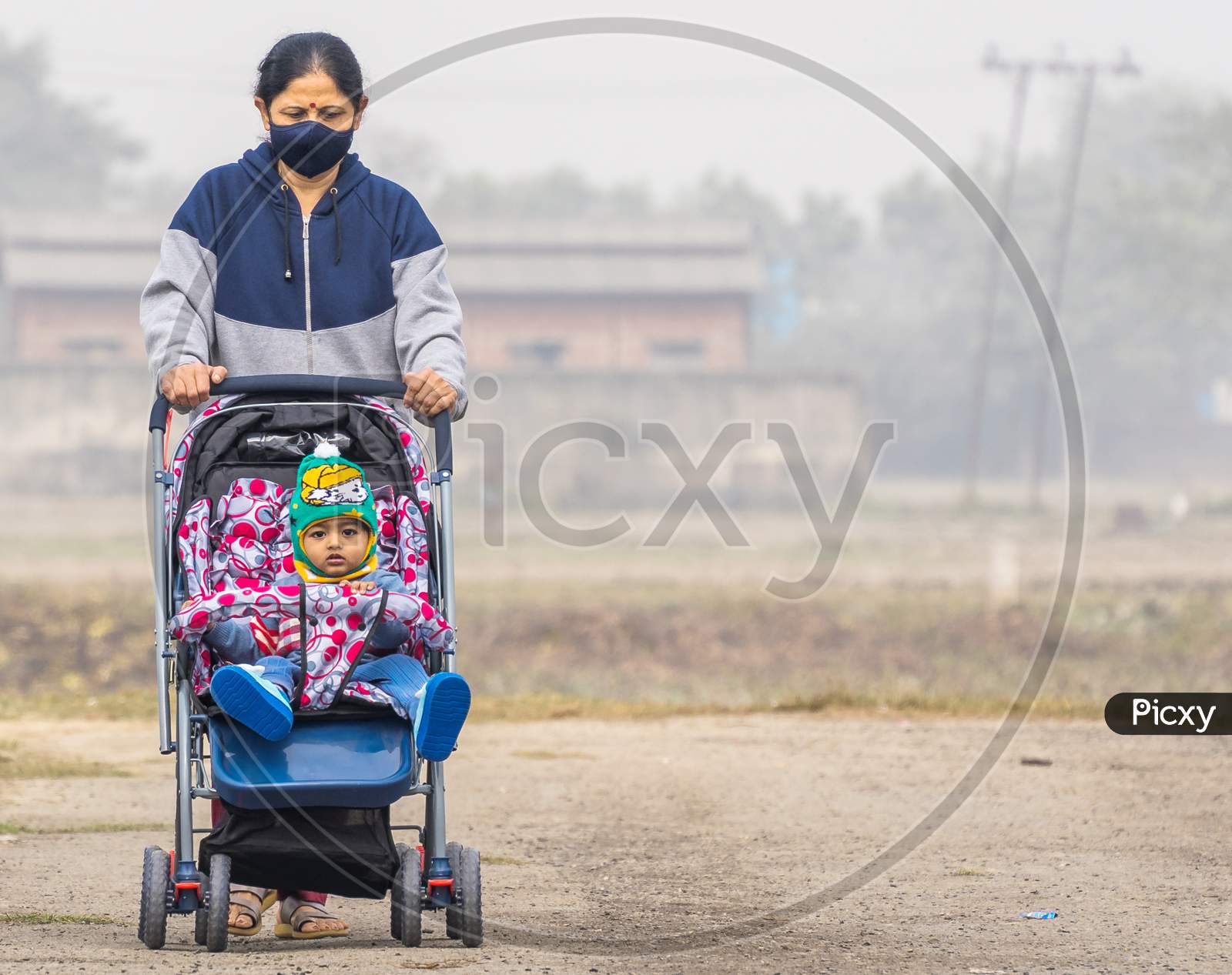 An Elderly Indian Lady Wearing Black Face Mask Pushing An Indian Baby In Winter Wear In A Stroller Perambulator On A Road With Copy Space And Background Blur