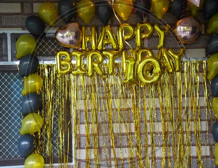 Golden Ribbons Or Happy Birthday Text Hanging On The Wall And Black And Gold Air Balloon Also Decorated On Wall.