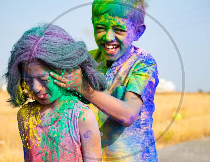 Young Kid From The Back Applying Holi Colors To Girls Face During Holi Festival Celebration - Concept Of Kids Having Fun By Playing Holi During Festive.