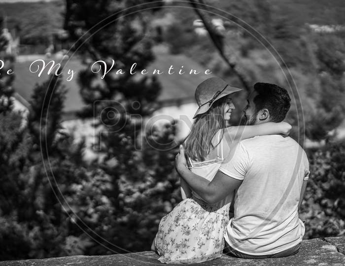 Be My Valentine. Couple Sitting On Stone Wall Embracing, Woman With Straw Hat Has A Smile On Face.