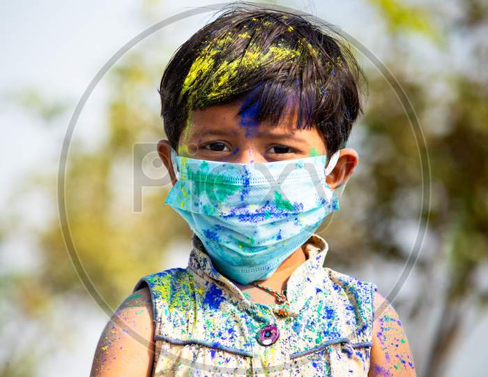 Portrait Of Young Cute Little Girl Kid With Medical Face Mask With Applied Holi Powder Looking At Camera - Concept Of Holi Celebration During Coronavirus Or Covid-19 Pandemic With Safety Measures.