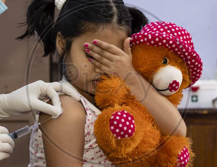 An Indian Girl Child With Teddy Bear Covers Her Eyes In Fear During Vaccination