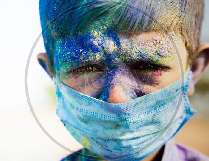 Head Shot Of Young Boy With Medical Face Mask Played Holi The Festival Of Colours And Looking Into Camera - Concept Of Holi Celebration During Covid-19 Coronavirus Pandemic.