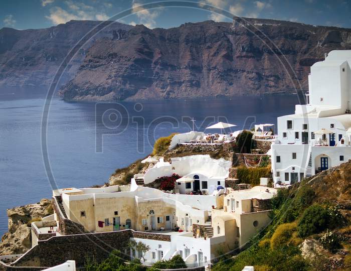 Santorini, Greece: Wide Angle Shot Of Cityscape Showing White Houses Built On Mountain Against Beach And Mountains During Summer