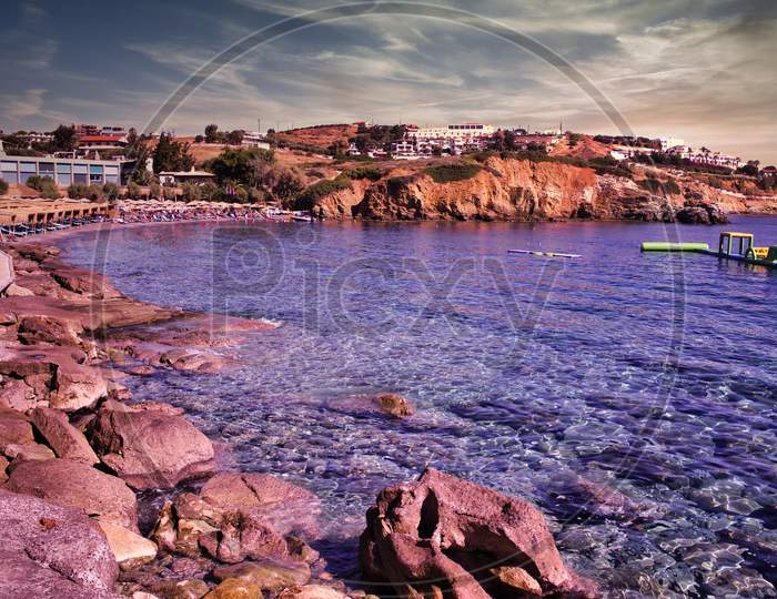 Crete Island, Greece: Wide Angle Shot Of Rocky Beach With Blue Sea Water With Luxury Swim Water Floats For Kids And Adults. Beach Cityscape Against Houses On Rocky Mountains