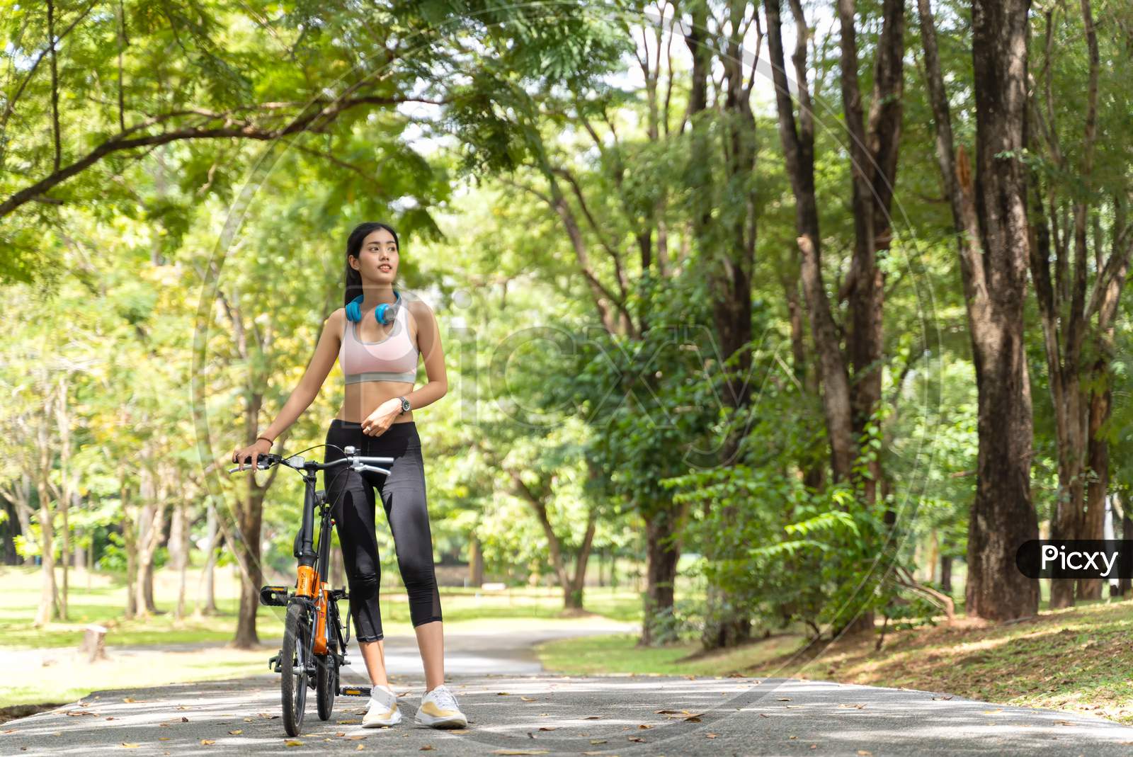 Healthy Asian Women With Headphones On The Neck Are Ride Bike In The Garden During Workout At The Park With Blur Bokeh Background In Sport And Healthy Concept With Copy Space.