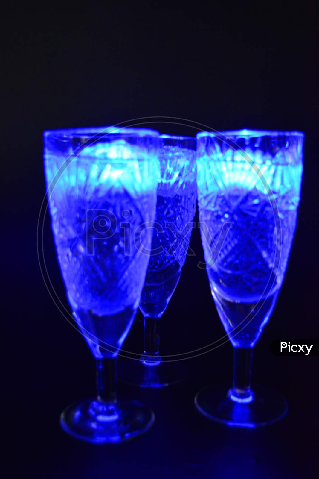Crystal champagne glasses with carvings in the form of snowflakes are located on a black matte background. Bright multicolor doidic glowing ice cubes float in the drink.