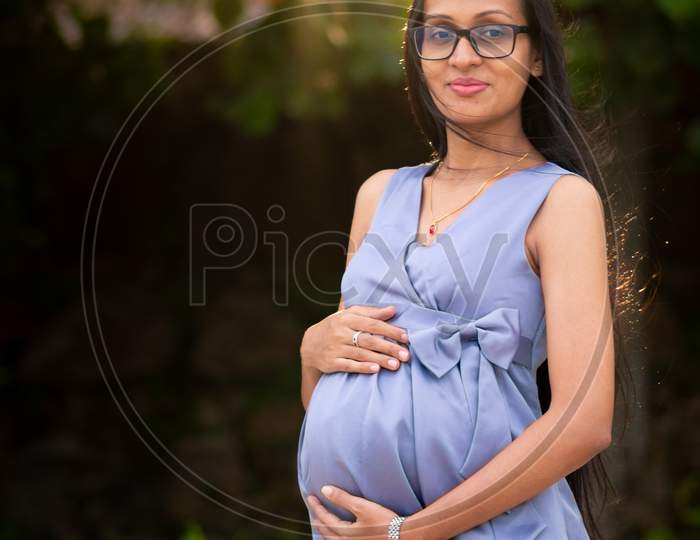 Young Adorable Looking Pregnant Lady Holding Belly With Both Hands Posing Outdoor, The Sun Setting Behind Her, Evening Light Shines Through Tree Leaves, Outdoor Photoshoot Of A Pregnant Woman In Galle