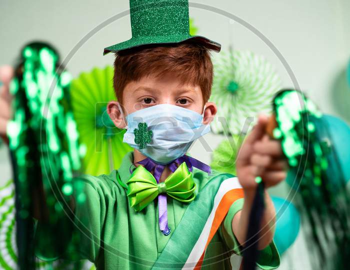 Young Kid With Medical Face Mask Dancing By Holding Pom Pom On Decorated Background At Home During Saint Patrics Day Celebration - Concept Of New Normal Festive Due To Coronvirus Covid-19 Pandemic.