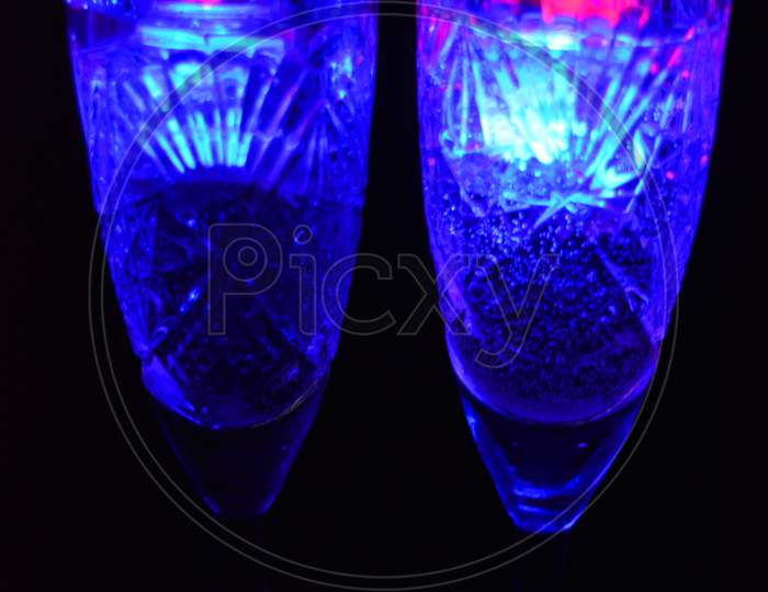 Crystal champagne glasses with carvings in the form of snowflakes are located on a black matte background. Bright multicolor doidic glowing ice cubes float in the drink.