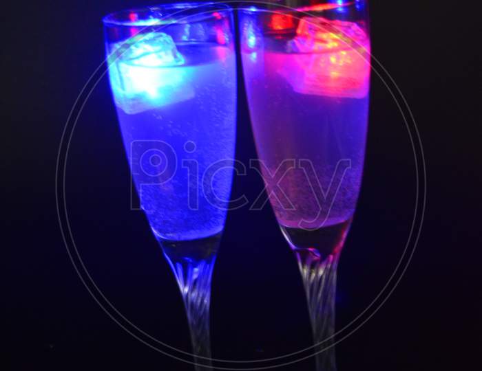 A champagne glass on a long swirling legs with a white drink is located on a black background. Luminous ice cubes of different colors with led diode illumination float in a glass.