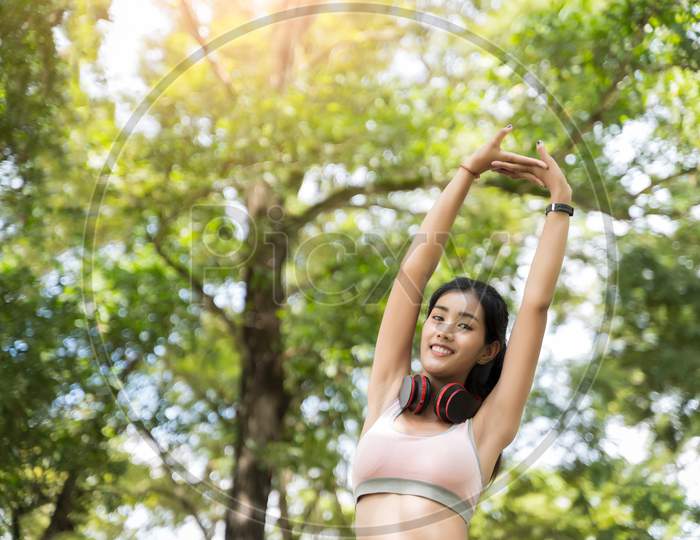 Healthy Asian Women In The Exchange Mood Are Stretching And Listening To Music With Headphones Before Starting A Workout At The Park With Blur Bokeh Background In Sport And Healthy Concept.