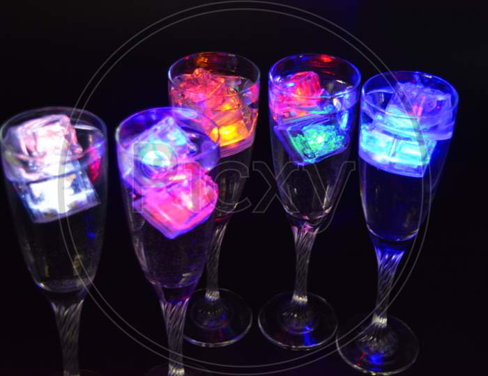 Five glass goblets on a long twisted legs with ice cubes glowing in different colors. Fun and bright drinks with luminous led elements are located on a black matte background, party and relaxation.