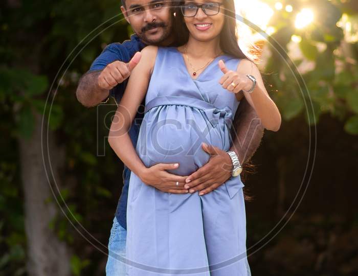 Mother To Be Wife Holding Her Belly By Both Hands, Father Stay Behind And Gives Thumbs-Up Gesture, Happy Couple Posing For A Photo Outdoor Evening Time, Couple Goals Concept,