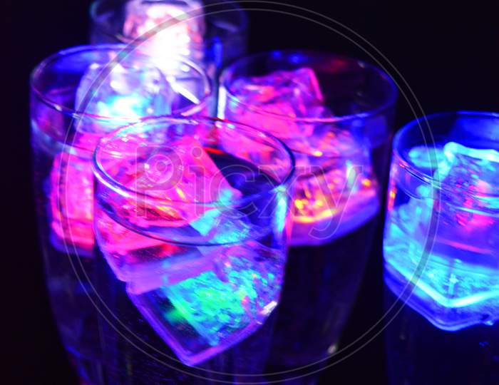 Five glass goblets on a long twisted legs with ice cubes glowing in different colors. Fun and bright drinks with luminous led elements are located on a black matte background, party and relaxation.
