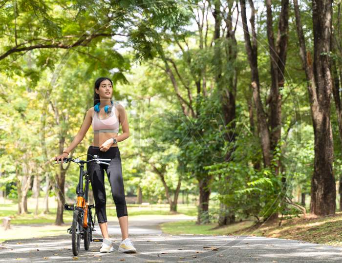 Healthy Asian Women With Headphones On The Neck Are Ride Bike In The Garden During Workout At The Park With Blur Bokeh Background In Sport And Healthy Concept With Copy Space.
