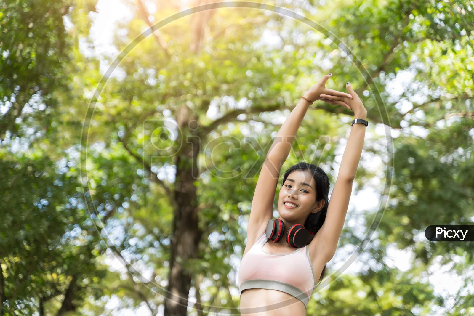 Healthy Asian Women In The Exchange Mood Are Stretching And Listening To Music With Headphones Before Starting A Workout At The Park With Blur Bokeh Background In Sport And Healthy Concept.